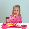 A young girl eating with matching pink dishes and utensils along with a pink Feeding Bowl made from Silicone with raisins inside.
