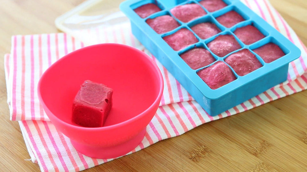 A pink Feeding Bowl made from Silicone with a frozen puree cube and sitting beside a blue silicone cube tray.