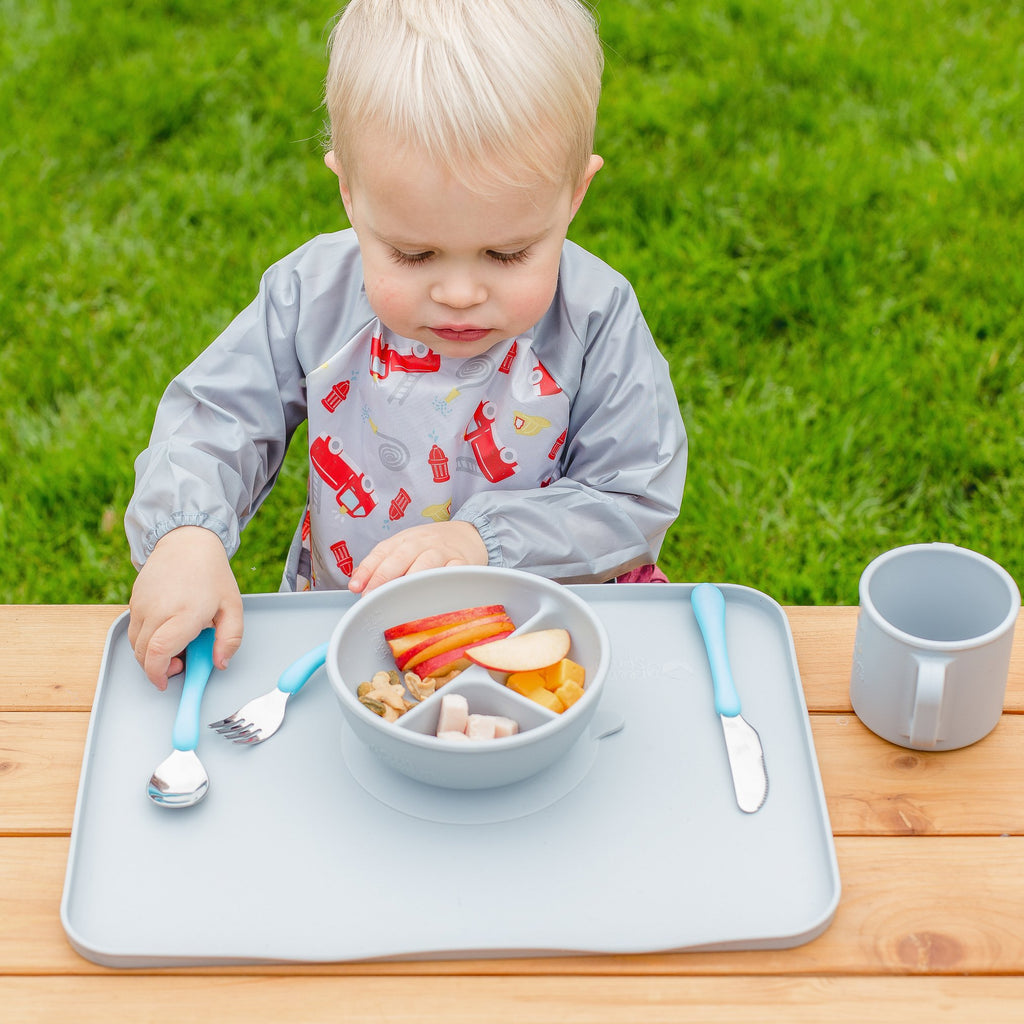 A young blond boy sitting outside looking at the food in the gray Learning Bowl made from Silicone