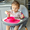 A little girl sitting in her high chair using her pink Learning Cutlery Set to get food from her pink bowl