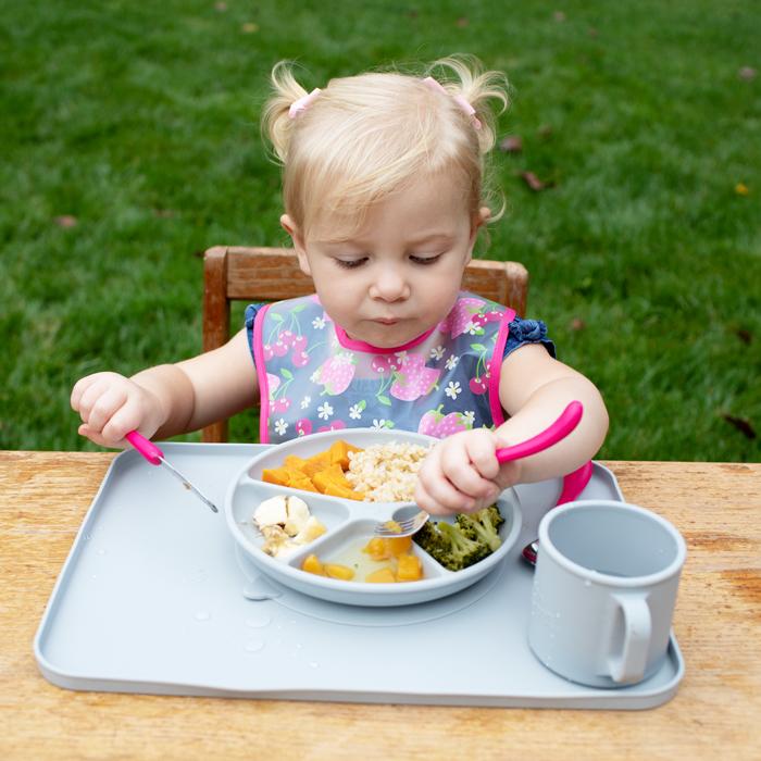 A young girl with pigtails sitting outside using the pink Learning Cutlery Set