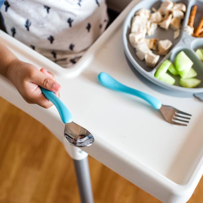 A little hand holding a aqua spoon from the Learning Cutlery Set while sitting the high chair
