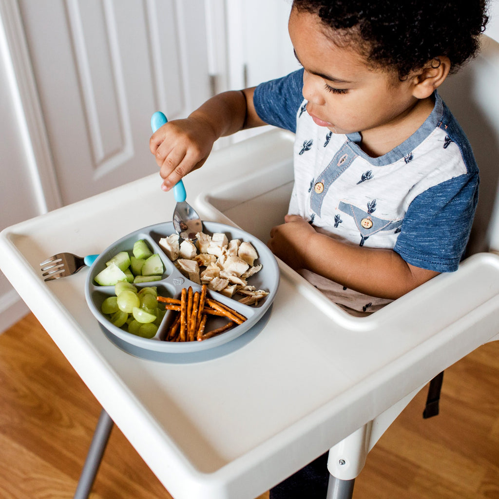 A young boy in a high chair using the aqua Learning Cutlery Set to eat his food