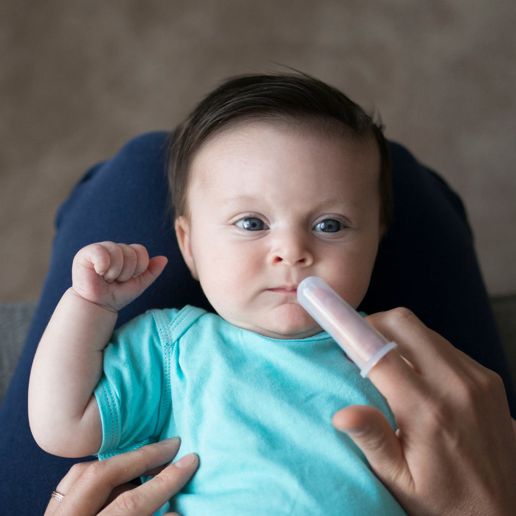 A mother with First Toothbrush made from Silicone on her finger is gently trying to coax her baby to open his mouth as he looks up at the viewer.