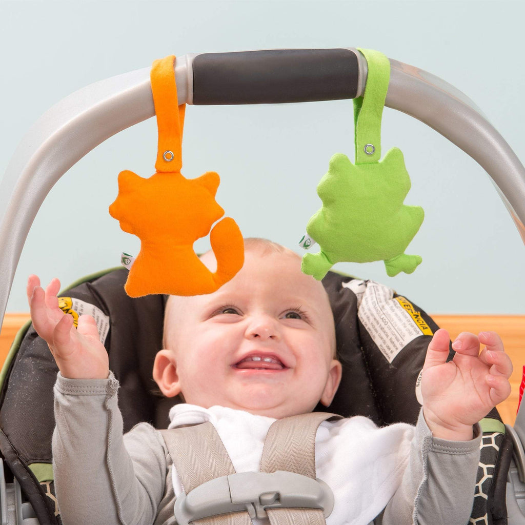 The orange fox and green frog friends are attached to the handle of a carrier as a smiling baby reaches up to play.