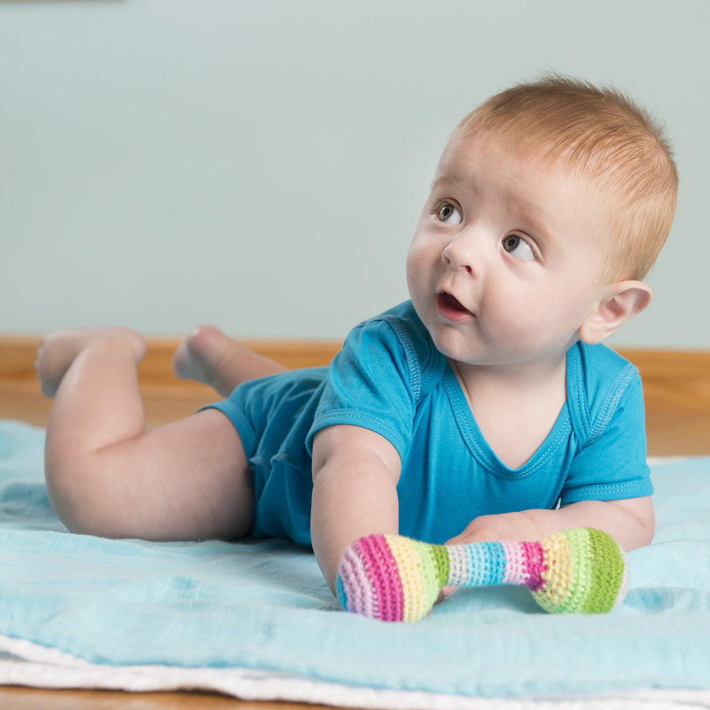 A baby boy laying on his stomach on a light blue blanket looking up at someone out of view with the Chime Rattle in front of him.