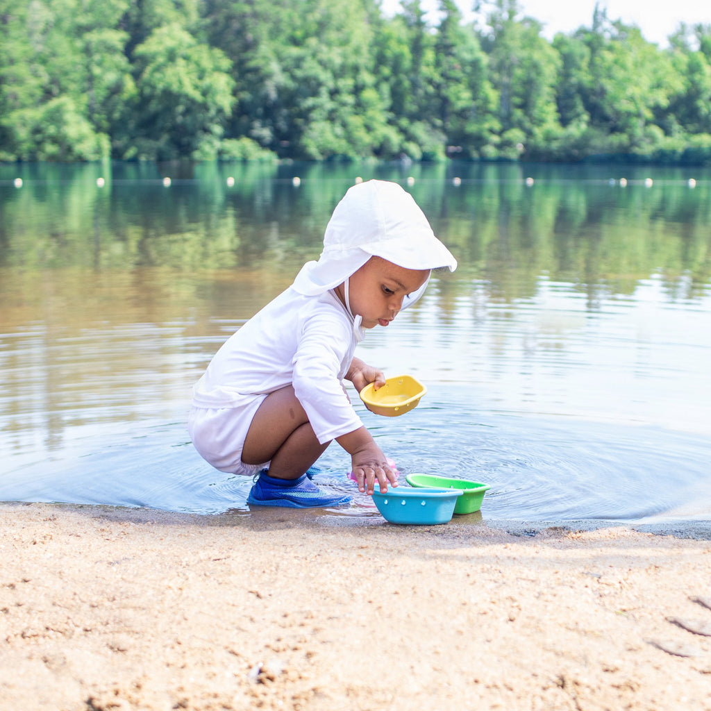 A toddle squatting on a beach to play with water boats in the lake while wearing an all white swim outfit along with a White Flap Sun Protection Hat.