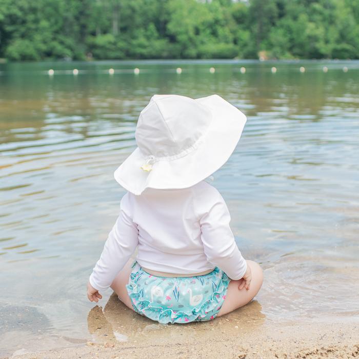 A baby girl facing away and sitting one a beach on lake while wearing a white Brim Sun Protection Hat.