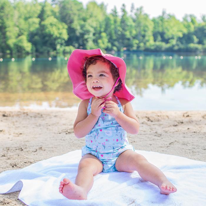 An adorable young girl wearing a one-piece swimsuit and a hot pink Brim Sun Protection Hat looking up while playing with the hat strings.