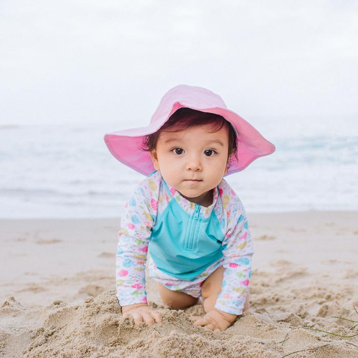A little baby girl crawling on the beach towards the viewer while wearing a light pink Brim Sun Protection Hat.