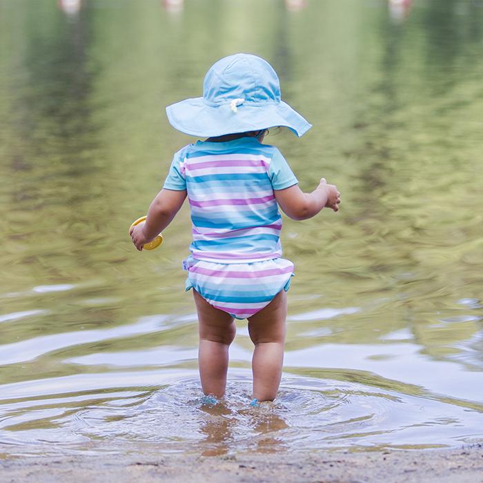 A toddler standing in the lake facing away while wearing a light blue Brim Sun Protection Hat.
