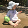 A little toddler looking to the right while sitting along a river holding a toy boat and wearing a gray Brim Sun Protection Hat.