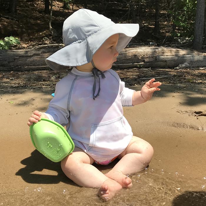 A little toddler looking to the right while sitting along a river holding a toy boat and wearing a gray Brim Sun Protection Hat.