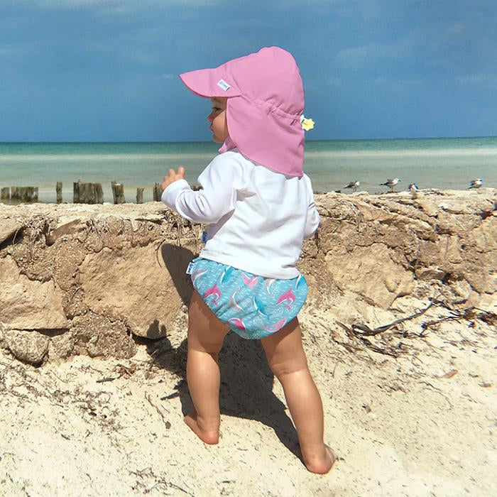 A young toddler looking to the side while standing next to a stone wall on a beach while wearing a light pink Flap Sun Protection Hat.