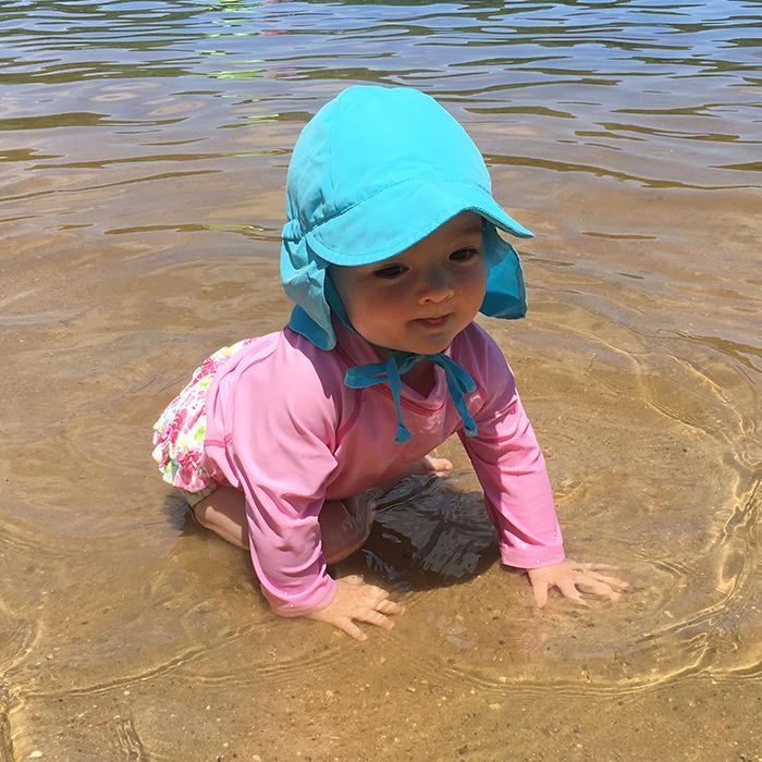 An adorable infant girl crawling through the water on the beach wearing a pink rashguard and an aqua Flap Sun Protection Hat.