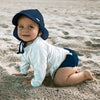 A cute toddler craddling along the beach while looking up and smiling and wearing a navy Flap Sun Protection Hat.