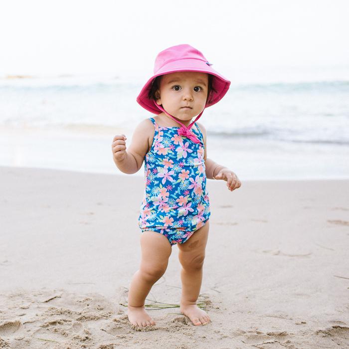 A young toddler girl trying to walk on the sand while wearing a hot pink Breathable Swim and Sun Bucket Hat.