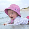 A smiling little girl laying on her belly in the shallow water on the beach while wearing an aqua Breathable Swim and Sun Bucket Hat.