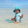 A little girl sitting in the shallow water on the beach while wearing an aqua Breathable Swim and Sun Bucket Hat.
