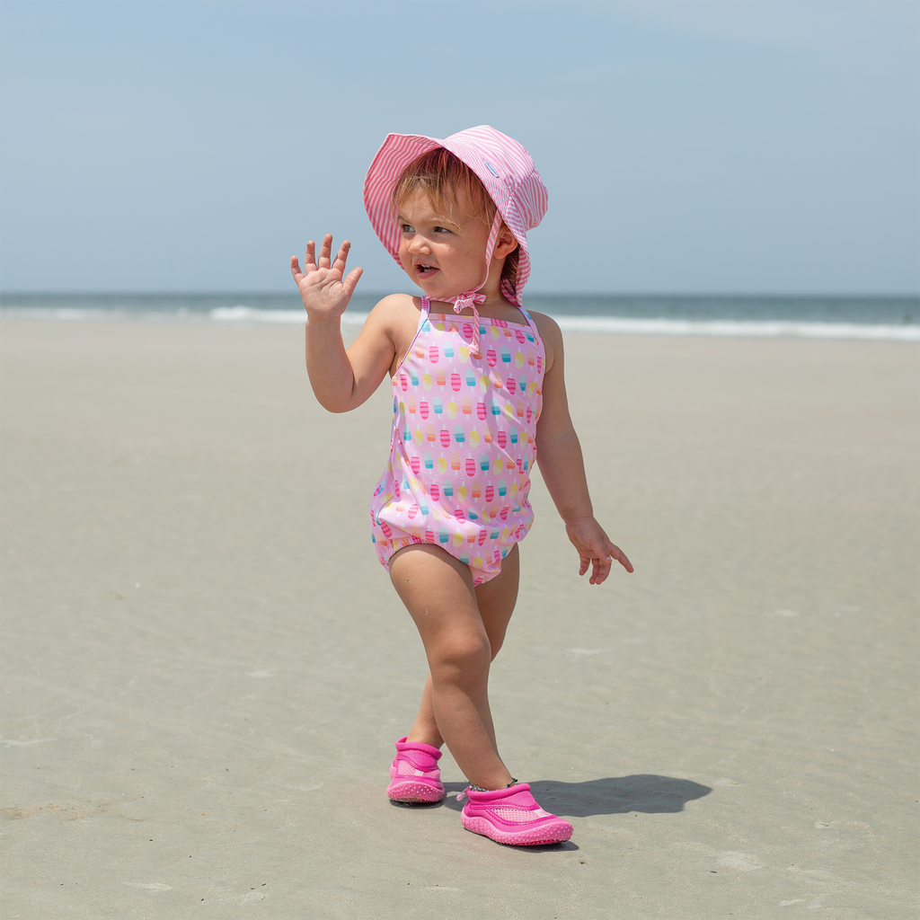 A young girl with a pink swim suit and the light pink pinstripe Bucket Sun Protection Hat walking on the beach waving toward someone behind her.
