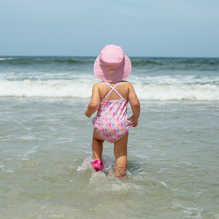 A young girl walking into the waves on the beach with a pink swim suit and the light pink pinstripe Bucket Sun Protection Hat.