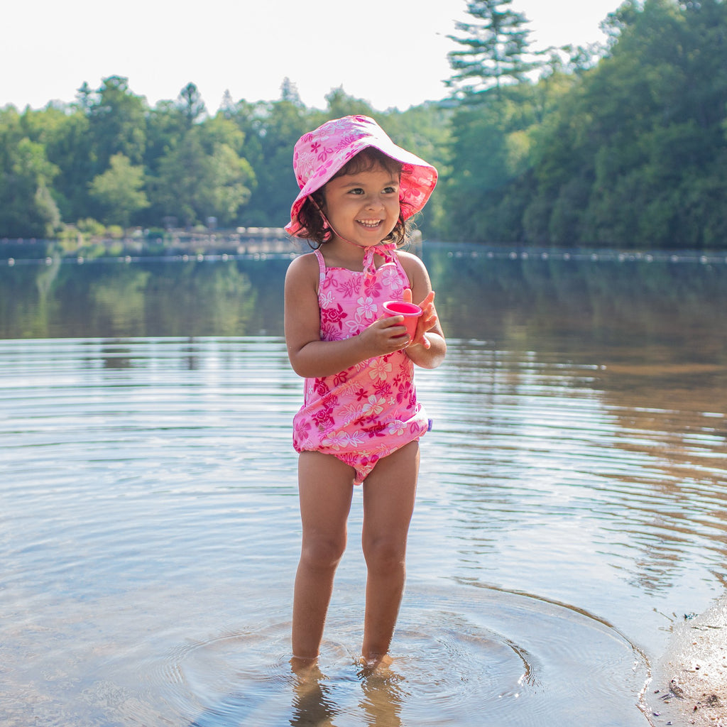 An adorable young girl standing in the shallow end of a lake holding a pink cup and wearing the pink hawaiian turtle Bucket Sun Protection Hat and a matching swimsuit.