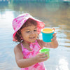 A young girl playing with toy water cups while wearing the pink hawaiian turtle Bucket Sun Protection Hat and a matching swimsuit.