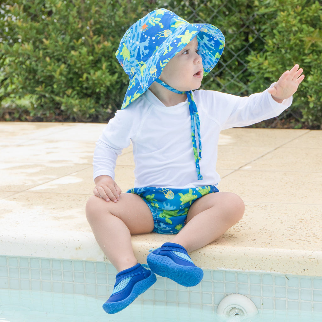 A young boy sitting on the edge of a pool in the backyard waving at someone out of view while wearing Royal Blue Turtle Journey Bucket Sun Protection Hat.