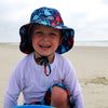 A happy young boy on a beach smiling at the camera while wearing a navy octopus Bucket Sun Protection Hat and white rashguard shirt.