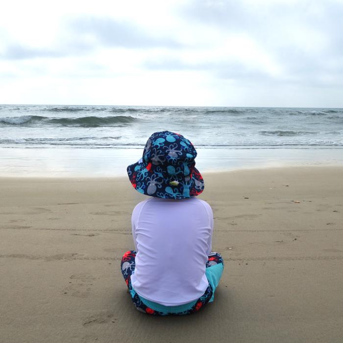 A young boy sitting along the beach looking out to sea while sporting the navy octopus Bucket Sun Protection Hat.