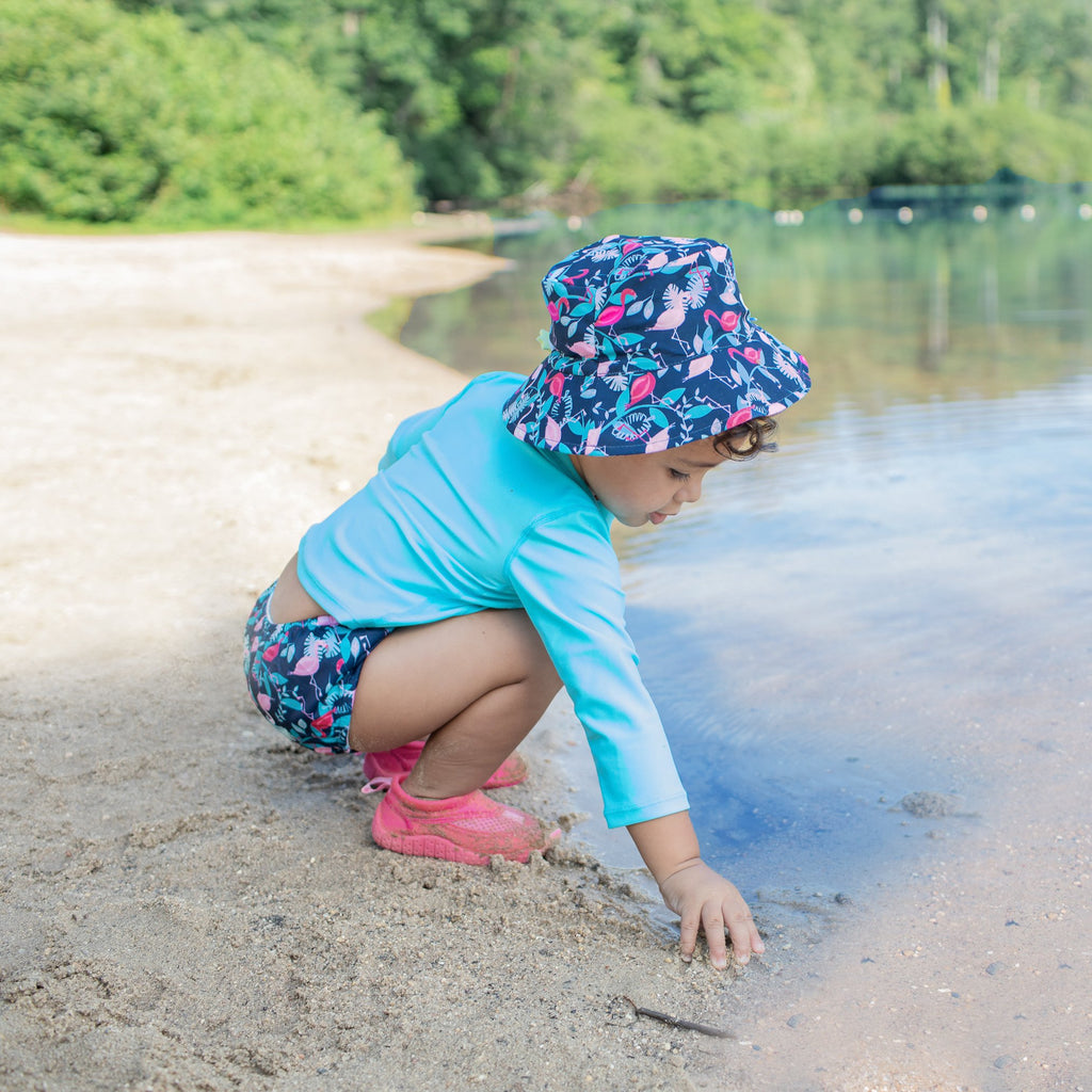 A young girl standing on the bank reaching toward the water while wearing the navy flamingo Bucket Sun Protection Hat.