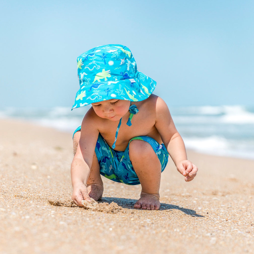 A cute toddler squatting and digging in the sand while wearing the Aqua Dinosaurs Bucket Sun Protection Hat and matching trunks.