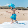 A toddler running through the waves on the beach while wearing the Aqua Dinosaurs Bucket Sun Protection Hat and matching trunks.