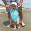 A smiling baby wearing the aqua Breathable Sun Protection Shirt while learning to walk and holding her parent's hand on the beach.