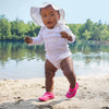 A little baby girl wabbling along the sand while wearing a white Brim Sun Protection Hat.