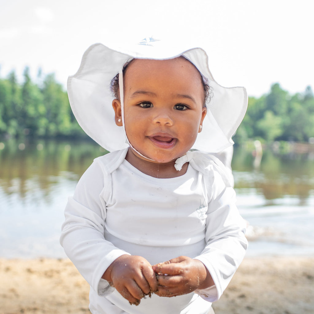 A pretty little baby girl with a white Brim Sun Protection Hat on looking into the camera while playing with sand in her hands.