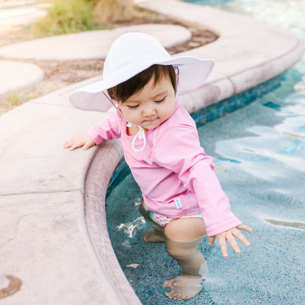 A little girl wearing a white Brim Sun Protection Hat standing in the pool while holding on to the edge and reaching intently at the water.