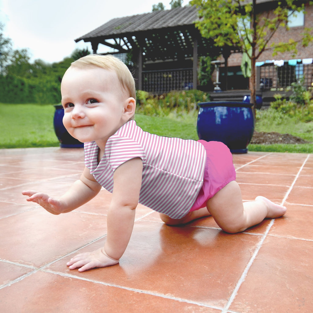A smiling young toddler girl crawling across tiled ground in the backyard while wearing a Light Pink Pinstripe Cap Sleeve Rashguard Shirt and pink swim diaper.