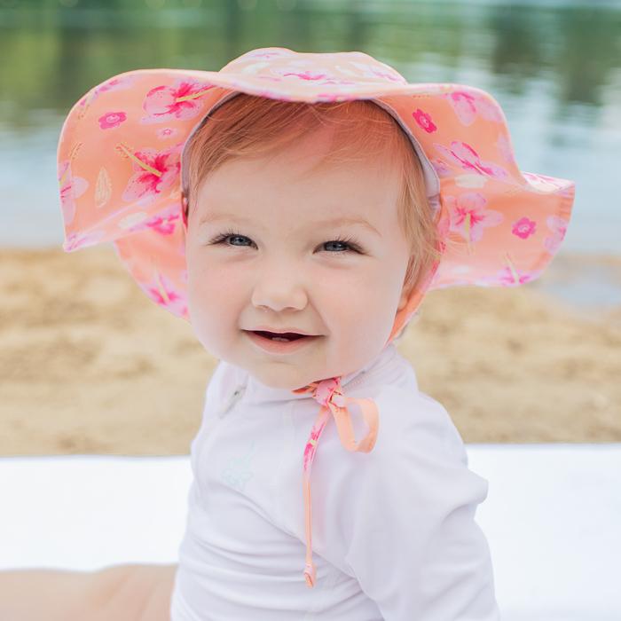 An adorable baby girl wearing the coral hibiscus Brim Sun Protection Hat while smiling up at the viewer.