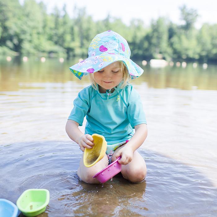 A little blonde girl kneeling in the shallow lake with an aqua coral reef Brim Sun Protection Hat while playing with toy boats.