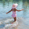 A cute young girl skipping away into the water towards the camera while wearing the light pink dragonfly floral Brim Sun Protection Hat and a matching swimsuit.