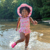 A cute young girl skipping through the water towards the camera while wearing the light pink dragonfly floral Brim Sun Protection Hat and a matching swimsuit. She has a funny cheesy smile going on.