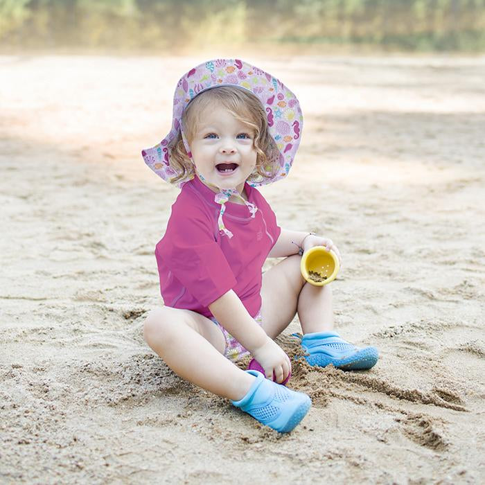A blonde curly haired girl wearing a pink sealife Brim Sun Protection Hat is playfully screaming while digging in the sand with toy cups.