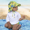 A toddler boy kneeling on his towel on the beach and wearing a Green Sealife Flap Sun Protection Hat. He is holding a sea shell but paying attention to something in the distance.
