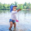 A young boy watching the water pour out of a toy boat he is holding up while standing in a lake and wearing the Royal Blue Sea Friends Flap Sun Protection Hat.